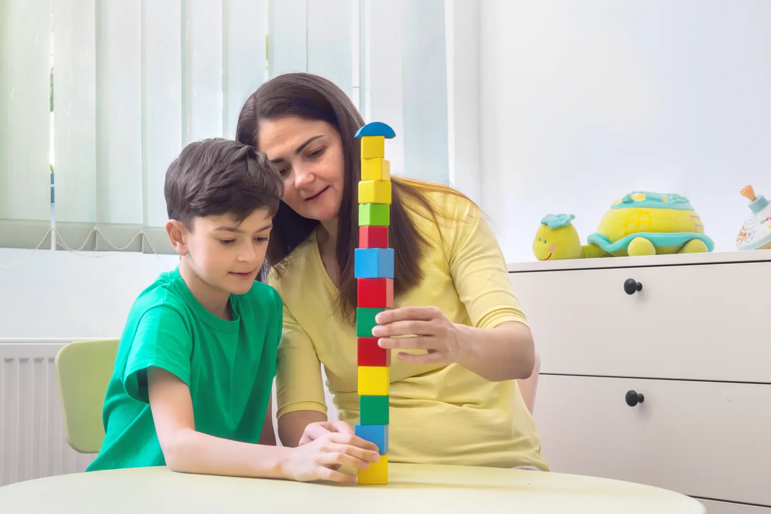 Praxis für Ergotherapie - medizinisch-therapeutische Behandlung - Kind und Therapeut bauen gemeinsam einen Turm aus Bausteinen während einer Entwicklungssitzung. Therapietechniken bei Autismus-Spektrum-Störung. Psychologische Unterstützung für Kinder. Interventionsaktivitäten bei Dyspraxie. Inklusiv